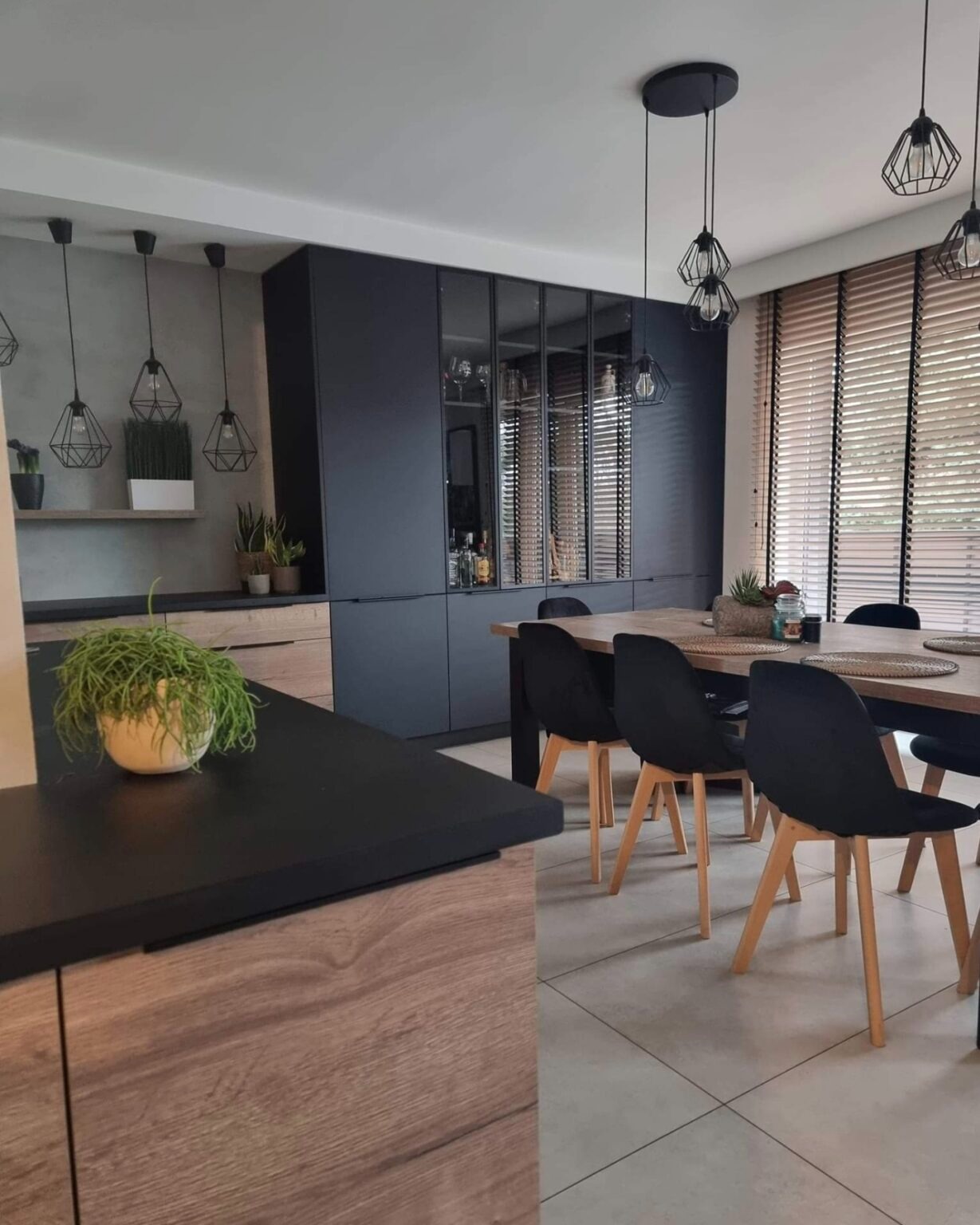 Nowoczesna kuchnia z czarnymi szafkami i drewnianymi akcentami, oświetlona wiszącymi lampami w stylu industrialnym, z widocznym stołem i krzesłami oraz roślinami doniczkowymi.