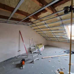 Poddasze w trakcie ocieplania wełną mineralną, widoczne metalowe profile sufitowe, drabina, poziomica, piła łańcuchowa i narzędzia na podłodze.