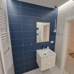 Nowoczesna łazienka z granatowymi płytkami, białym grzejnikiem drabinkowym i szafką pod umywalką w kremowym odcieniu, widoczna szklana kabina prysznicowa.