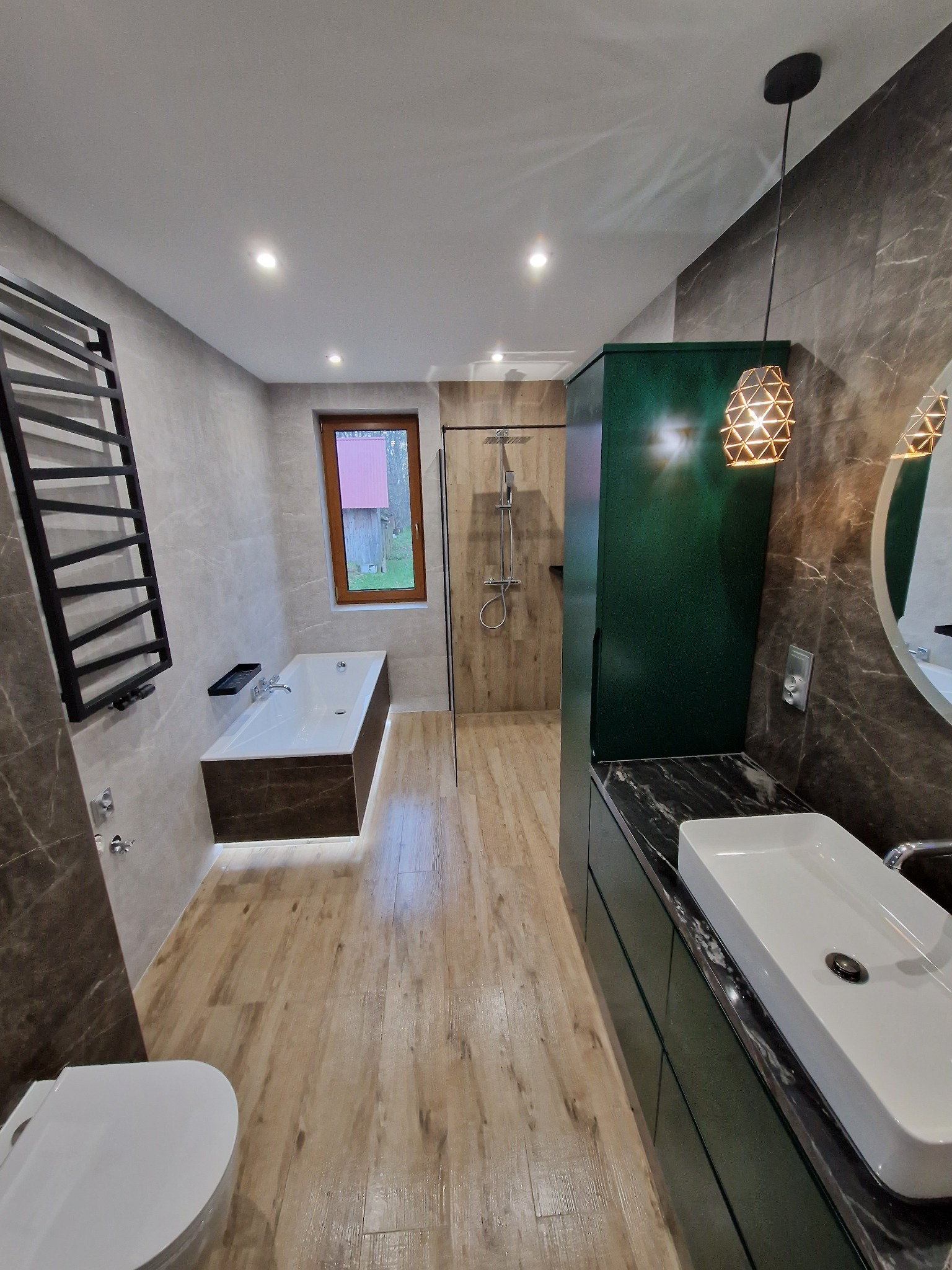 Nowoczesna łazienka z drewnopodobną podłogą, grafitowymi płytkami imitującymi marmur, wanną z podświetleniem LED, kabiną prysznicową walk-in, zieloną szafką z czarnym blatem i geometryczną lampą...