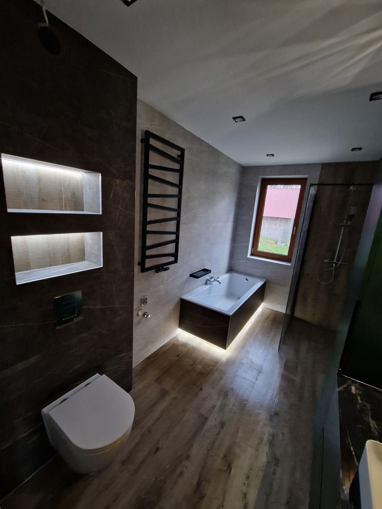 Nowoczesna łazienka z podświetlaną wanną, brązowymi płytkami imitującymi marmur, grzejnikiem drabinkowym i drewnopodobną podłogą, widok na zewnątrz przez okno.