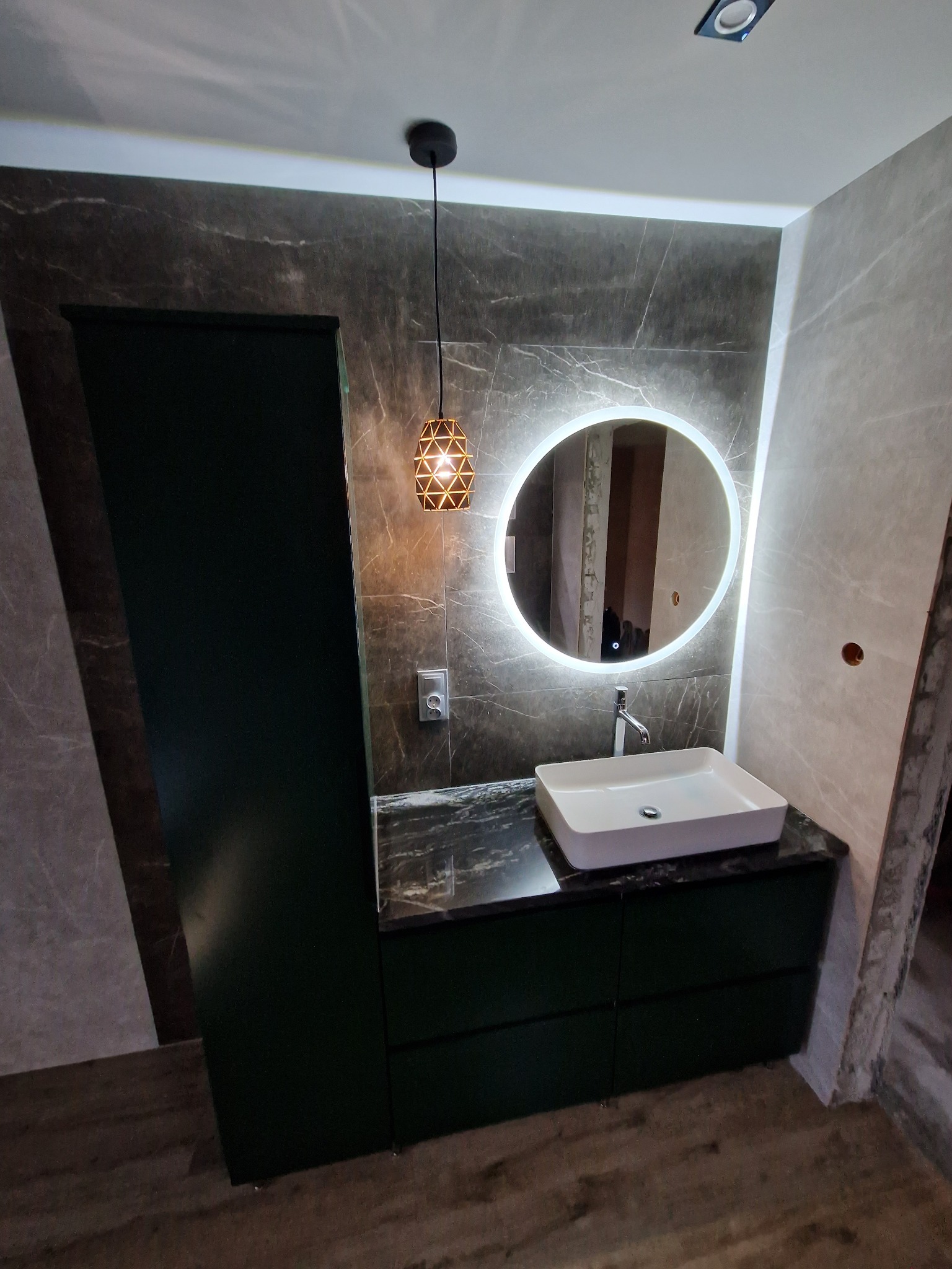 Nowoczesna łazienka z marmurowymi płytkami, okrągłym lustrem z podświetleniem LED, białą umywalką nablatową i ciemnozielonymi szafkami, oświetlona wiszącą lampą geometryczną.