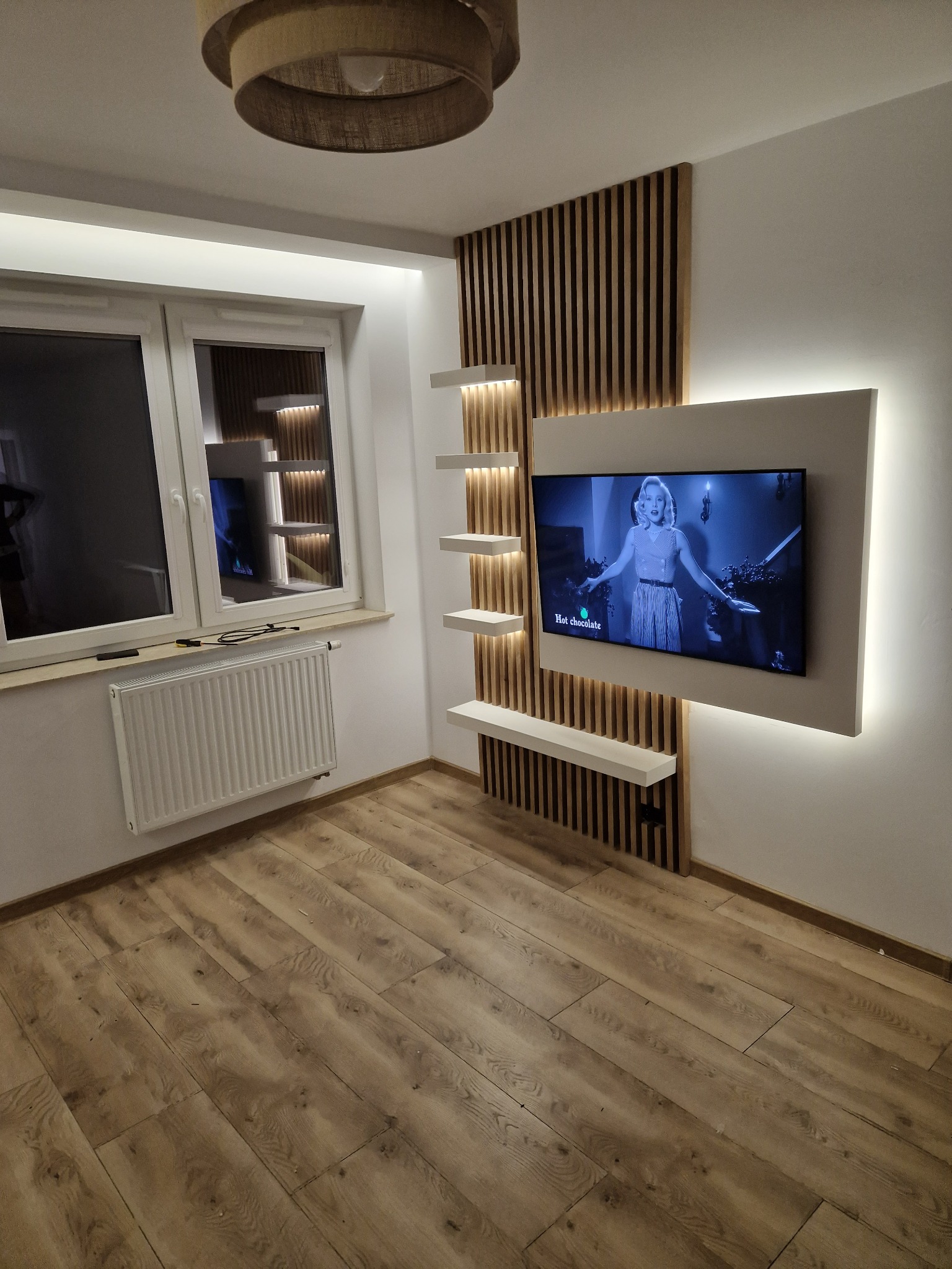 Nowoczesny salon z drewnianą ścianką działową, podświetlanymi półkami i telewizorem z czarno-białym filmem, oświetlenie sufitowe i panele podłogowe w ciepłych odcieniach brązu.