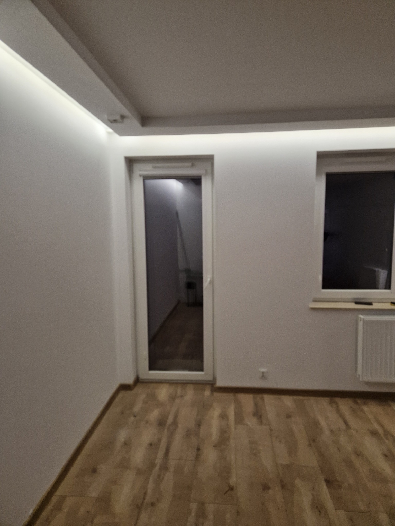 Pomalowane na biało wnętrze pokoju z drewnopodobną podłogą, białymi drzwiami i oknem, oświetleniem LED podsufitowym.