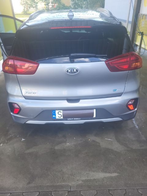 Tył srebrnego samochodu Kia Niro z otwartymi drzwiami, widoczny na myjni samochodowej z mokrą nawierzchnią.