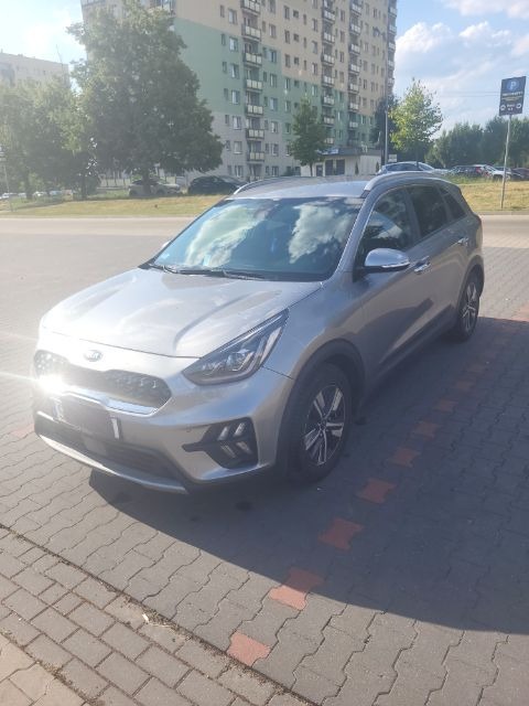 Srebrny samochód Kia Niro zaparkowany na chodniku z kostki brukowej, na tle bloku mieszkalnego i drzew.