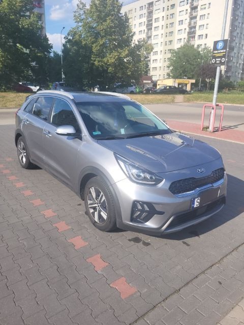 Szary samochód osobowy Kia Niro zaparkowany na kostce brukowej z elementami czerwonej kostki, na tle bloku mieszkalnego i drzew.