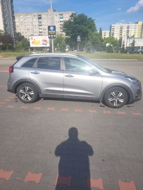 Szary samochód typu crossover zaparkowany na parkingu z czerwoną kostką, z widocznym cieniem osoby robiącej zdjęcie, w tle bloki mieszkalne i reklama parkingu.