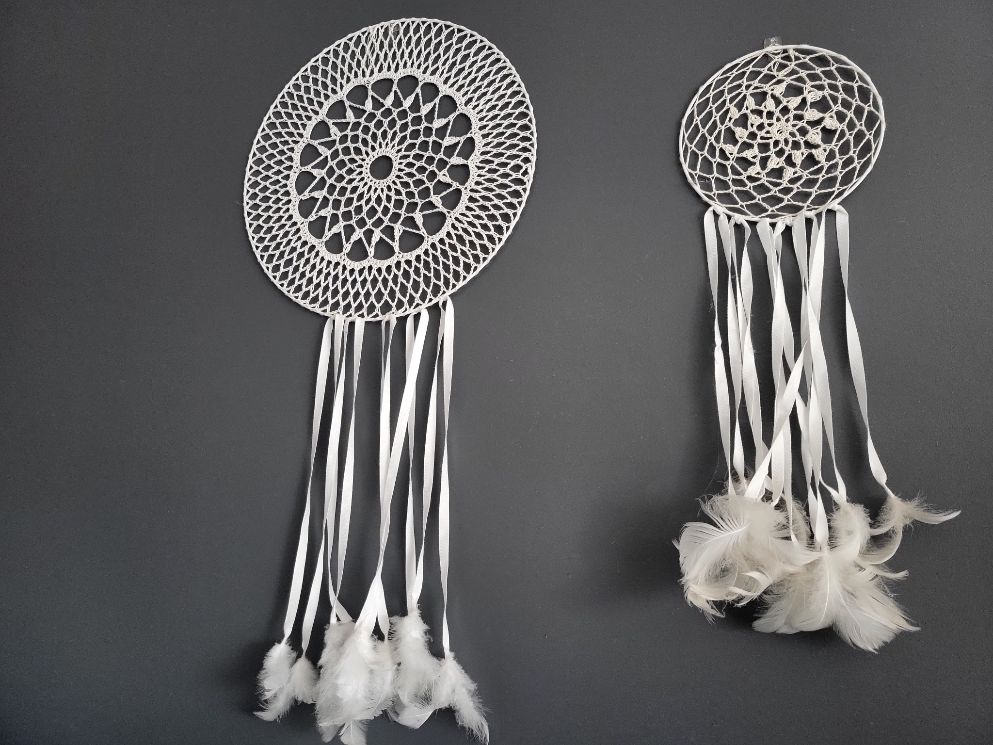 Dwa ręcznie robione łapacze snów z białej koronki i piór, zawieszone na szarej ścianie.