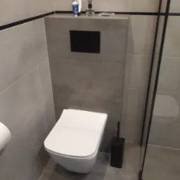 Nowoczesna łazienka z podwieszaną toaletą, szarymi płytkami, czarnymi akcentami i kosmetykami na zabudowie stelaża.