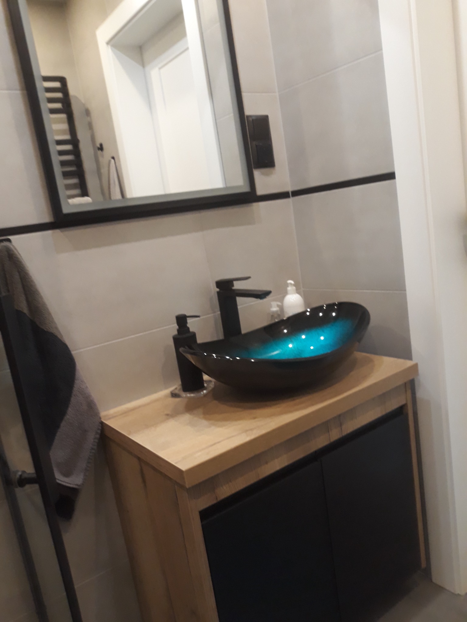 Nowoczesna łazienka z umywalką w kształcie misy, czarną armaturą i szafką z drewnianym blatem, widok na lustro i grzejnik ręcznikowy.