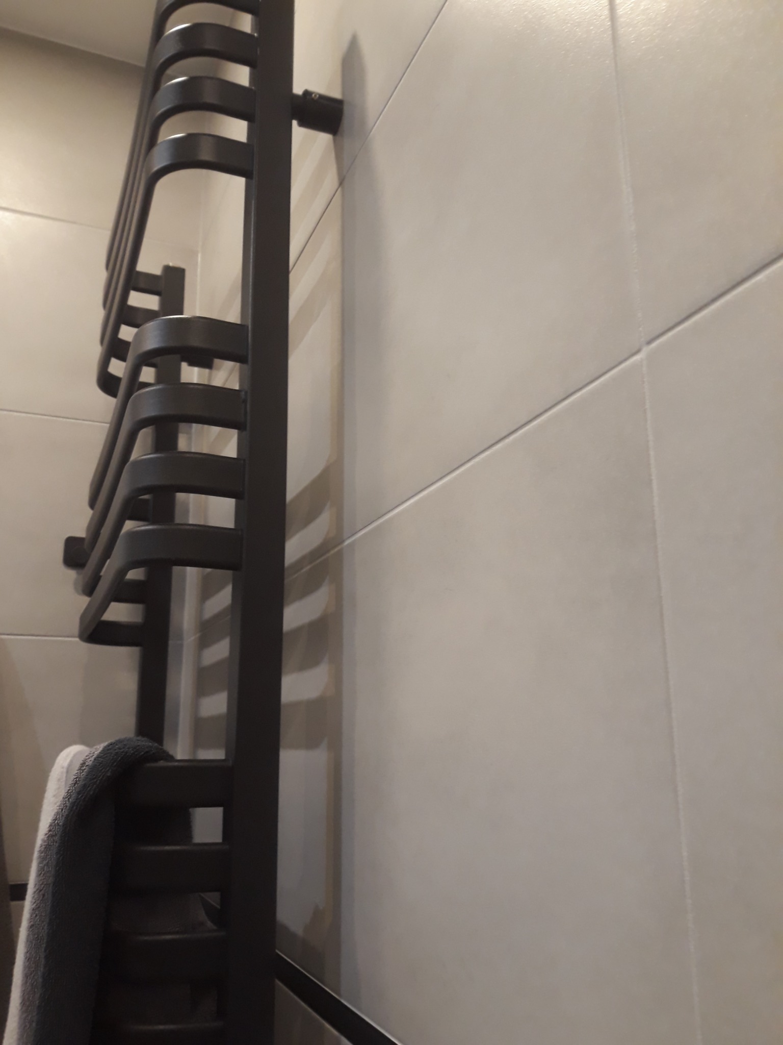 Czarny, drabinkowy grzejnik łazienkowy z szarym ręcznikiem, zamontowany na ścianie wyłożonej jasnymi, kwadratowymi płytkami ceramicznymi z delikatnym połyskiem.