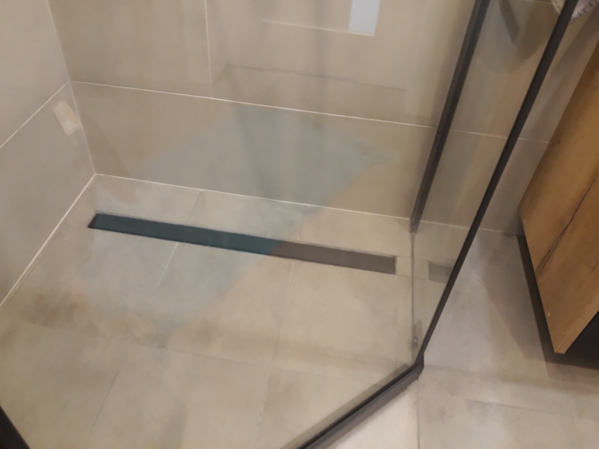 Beżowe płytki podłogowe i ścienne w kabinie prysznicowej z odpływem liniowym w ciemnej obudowie.