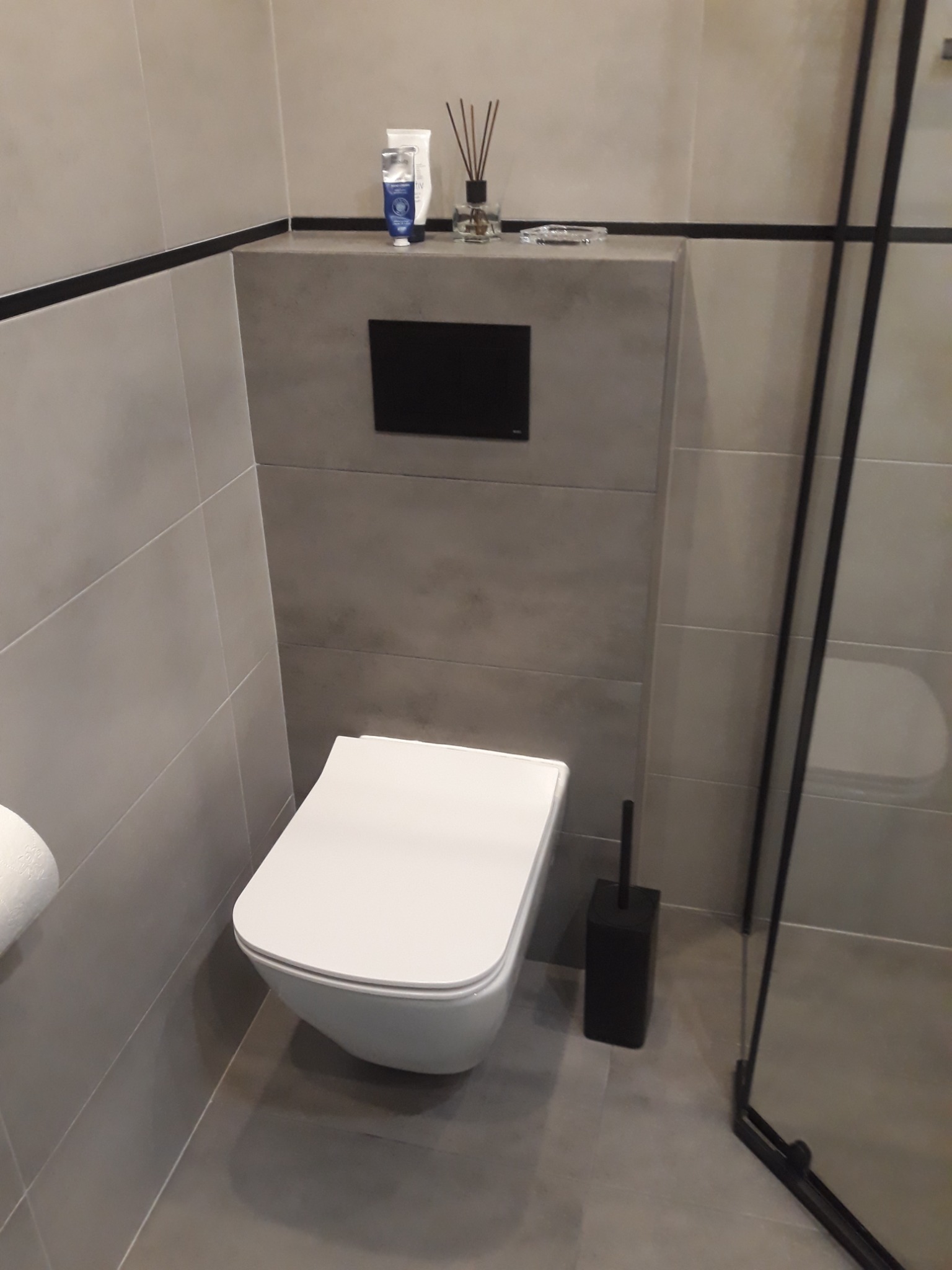 Nowoczesna łazienka z podwieszaną toaletą, szarymi płytkami, czarnymi akcentami i kosmetykami na zabudowie stelaża.