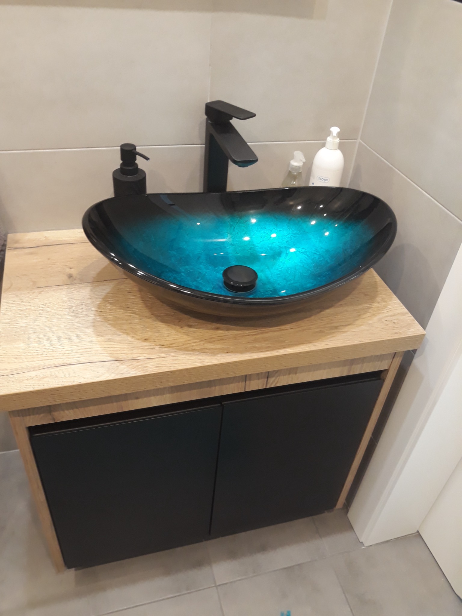 Błękitna, szklana umywalka nablatowa z czarną armaturą na szafce z drewnianym blatem i czarnymi frontami w nowoczesnej łazience.
