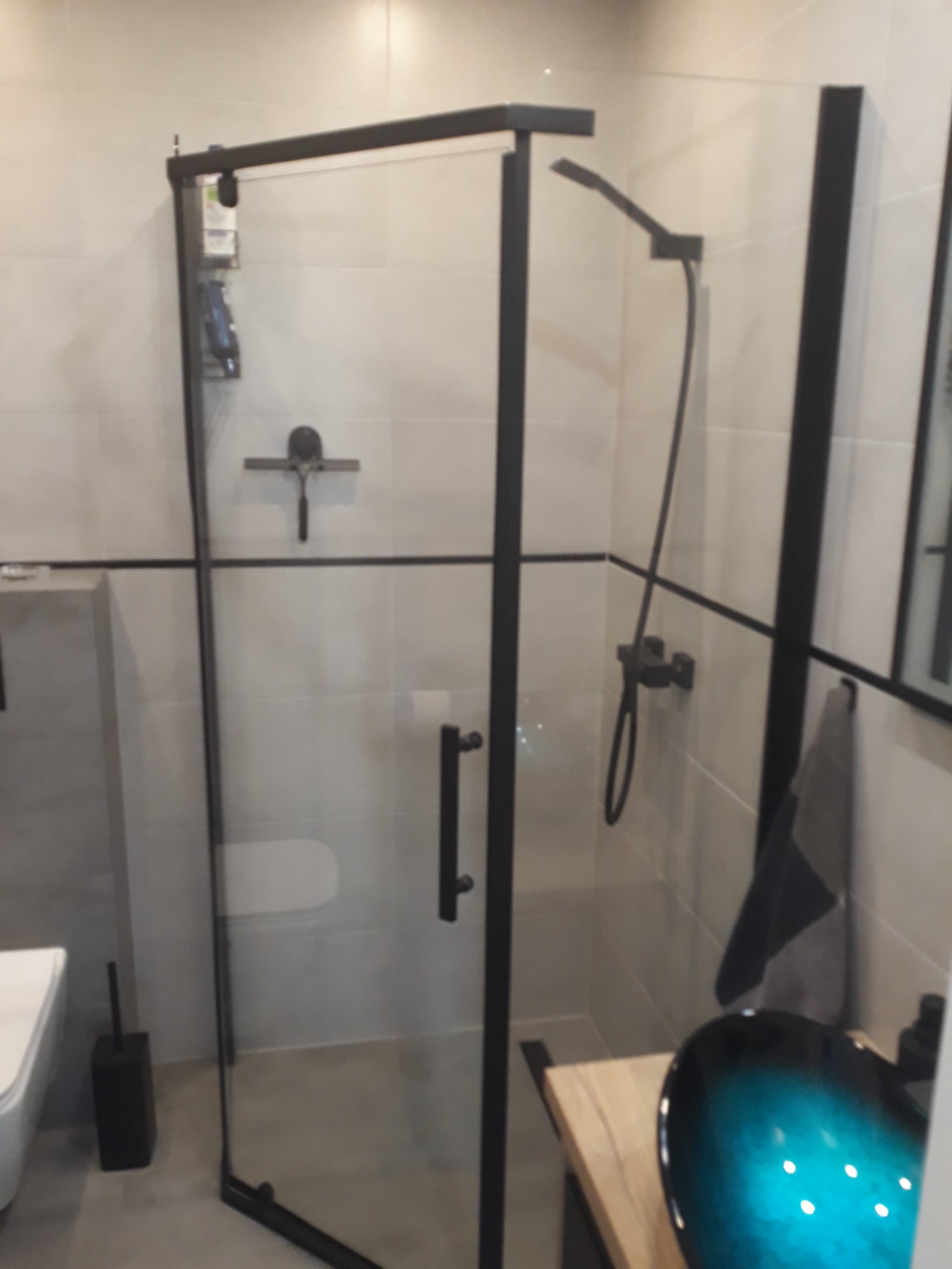 Nowoczesna łazienka z kabiną prysznicową typu walk-in z czarnymi profilami, deszczownicą i umywalką w kolorze turkusowym na drewnianym blacie.