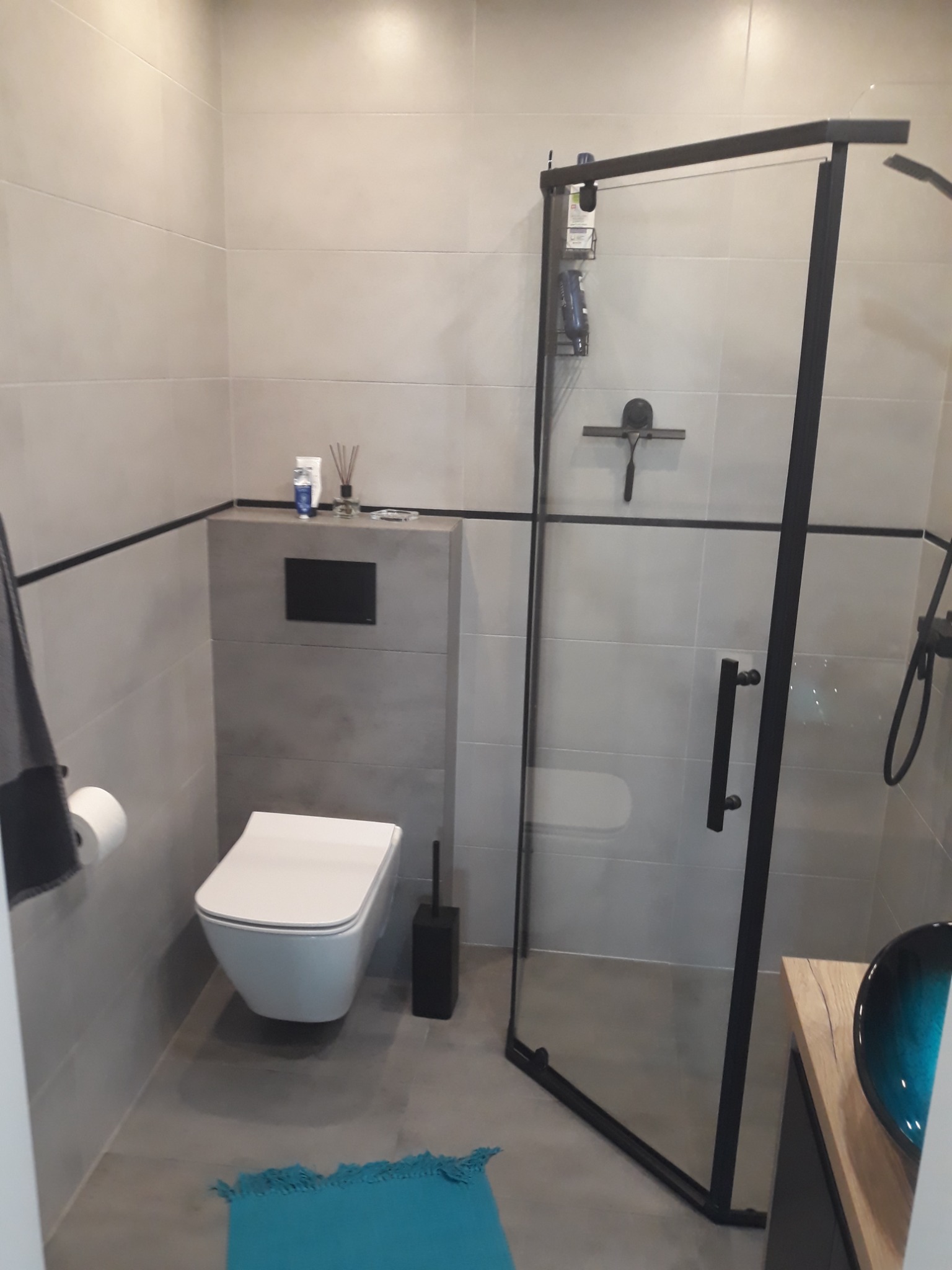 Nowoczesna łazienka z szarą glazurą, kabiną prysznicową z czarnymi ramami, podwieszaną toaletą, turkusowym dywanikiem i drewnianą szafką z umywalką w odcieniach niebieskiego.