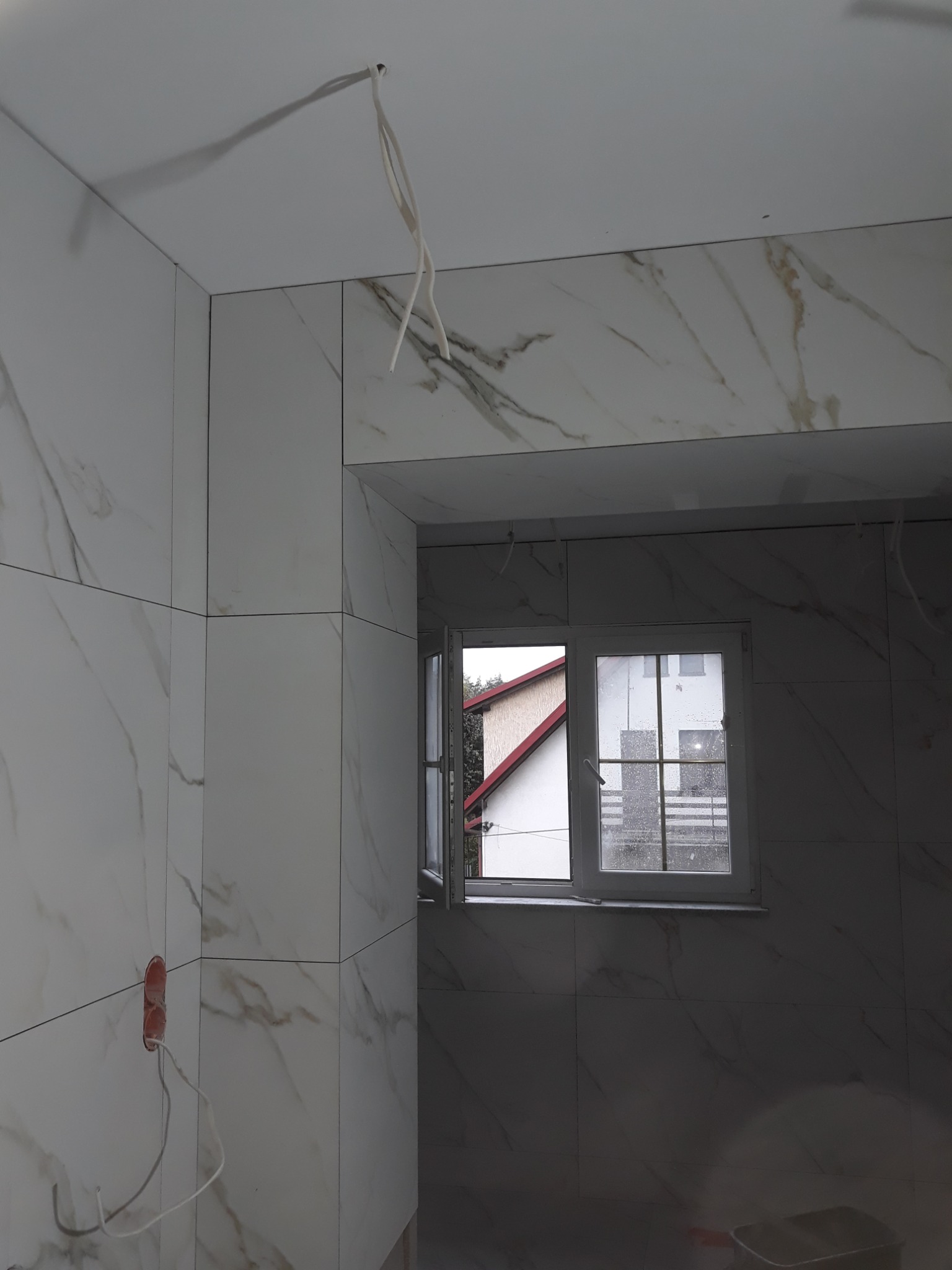 Łazienka w trakcie remontu: ściany i sufit wyłożone białymi płytkami imitującymi marmur, widoczne kable wystające z sufitu i puszki elektrycznej, okno z widokiem na dom.