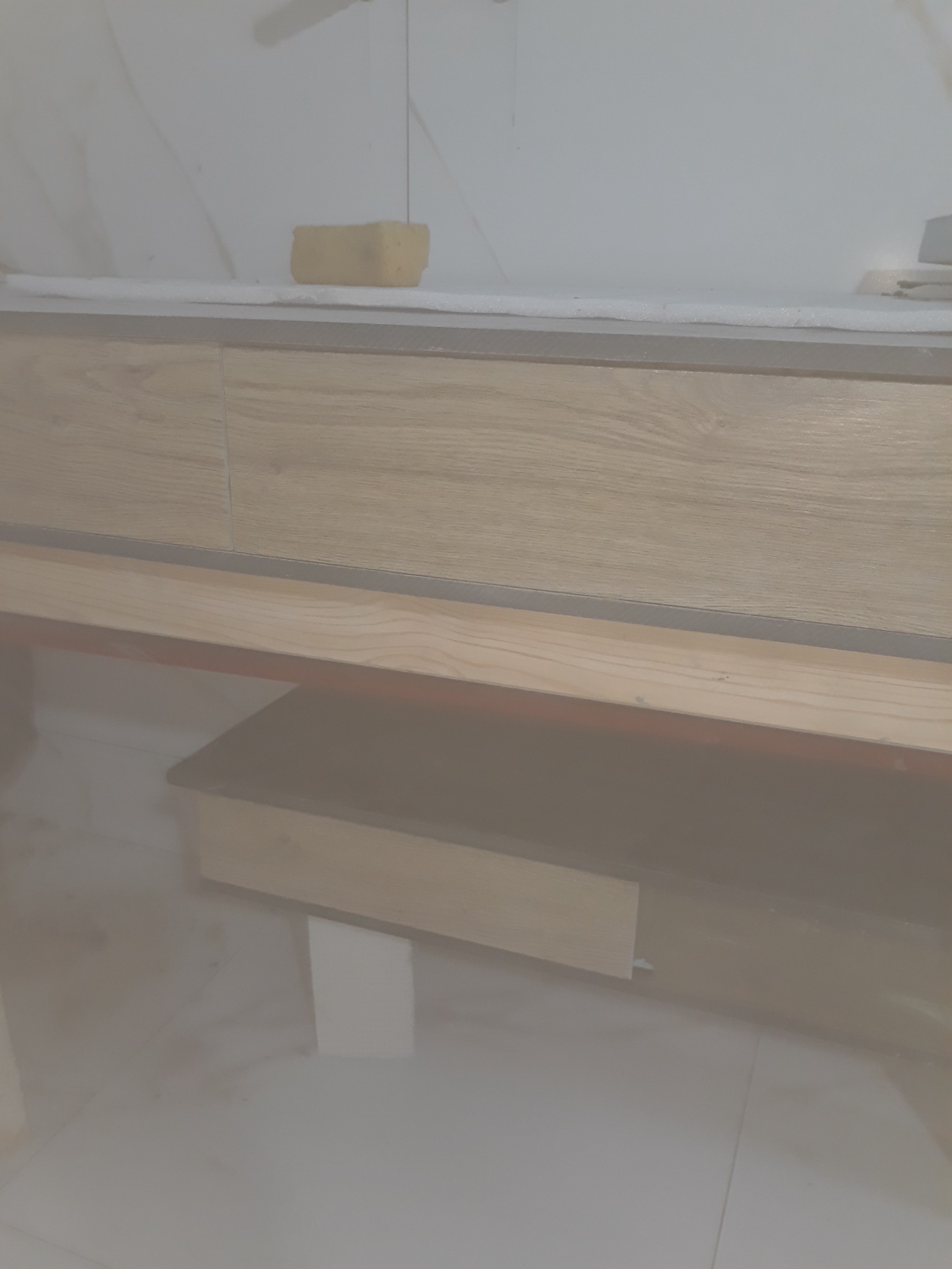 Montaż szafki pod umywalkę z drewnianymi frontami, widoczna gąbka i biała piana na blacie, marmurowe płytki podłogowe.