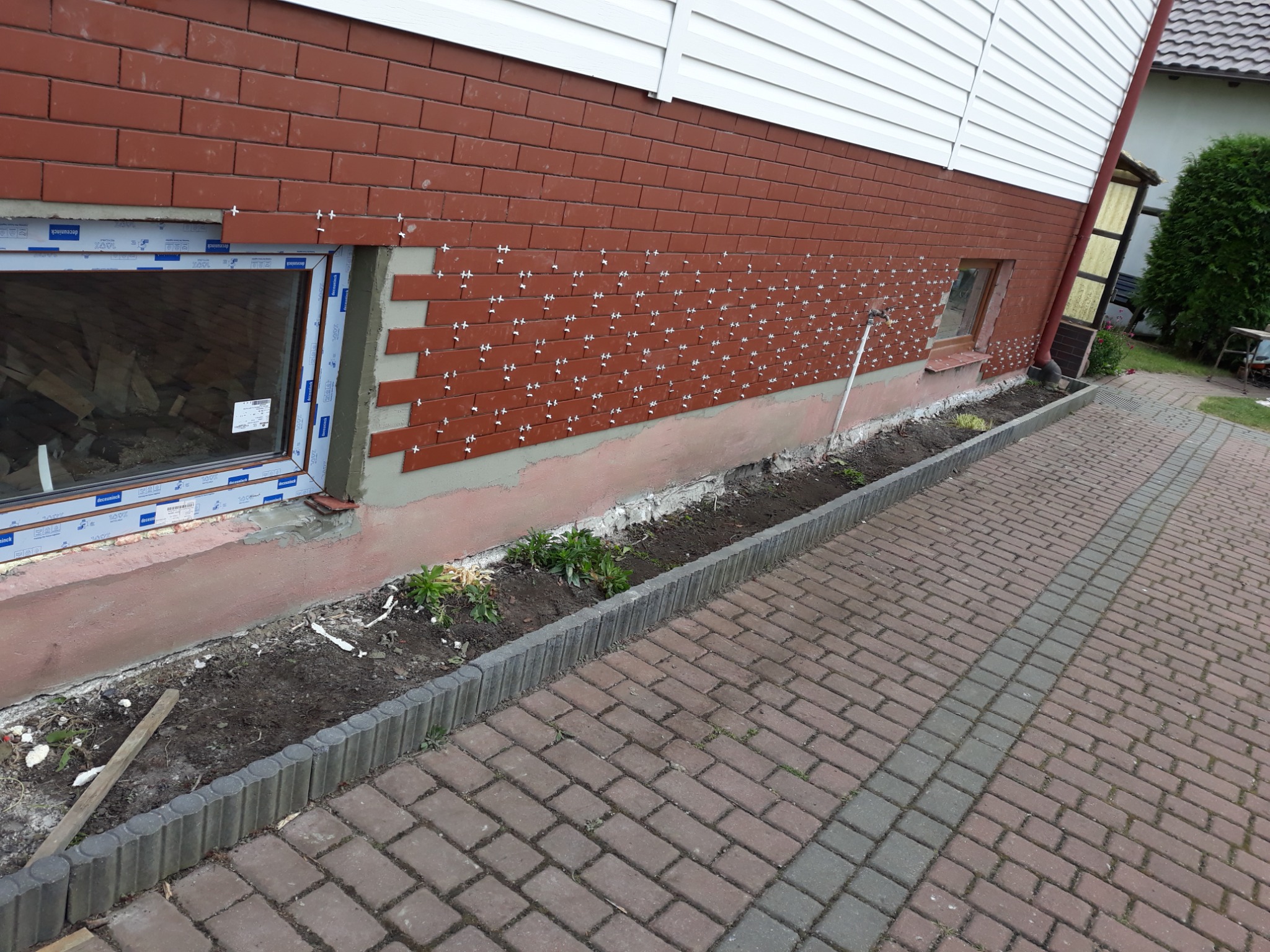 Montaż cegieł klinkierowych na elewacji budynku, widoczne krzyżyki dystansowe, okno z folią ochronną i fragment chodnika z kostki brukowej.