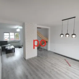 Jasne, przestronne wnętrze po remoncie z szarymi panelami podłogowymi, białą cegłą dekoracyjną i nowoczesnym oświetleniem wiszącym.