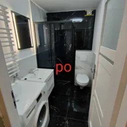 Nowoczesna łazienka z czarnymi marmurowymi płytkami, białą armaturą i pralką Bosch, oświetlona lampą sufitową z efektem kryształu.