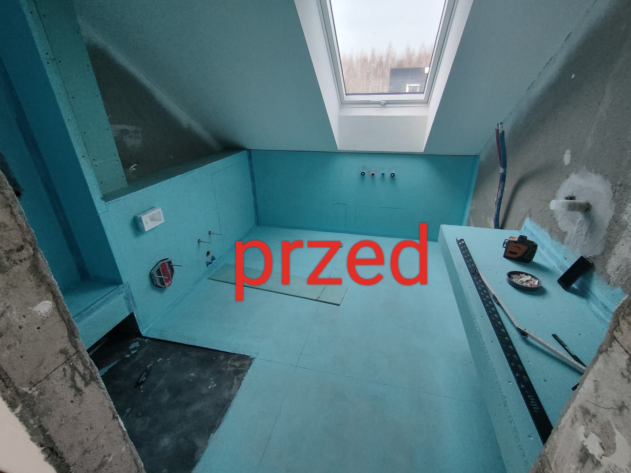 Łazienka na poddaszu w trakcie przygotowania do remontu: ściany i podłoga pokryte niebieskimi płytami izolacyjnymi, widoczne przewody instalacji, okno dachowe, laser krzyżowy i listwa pomiarowa.