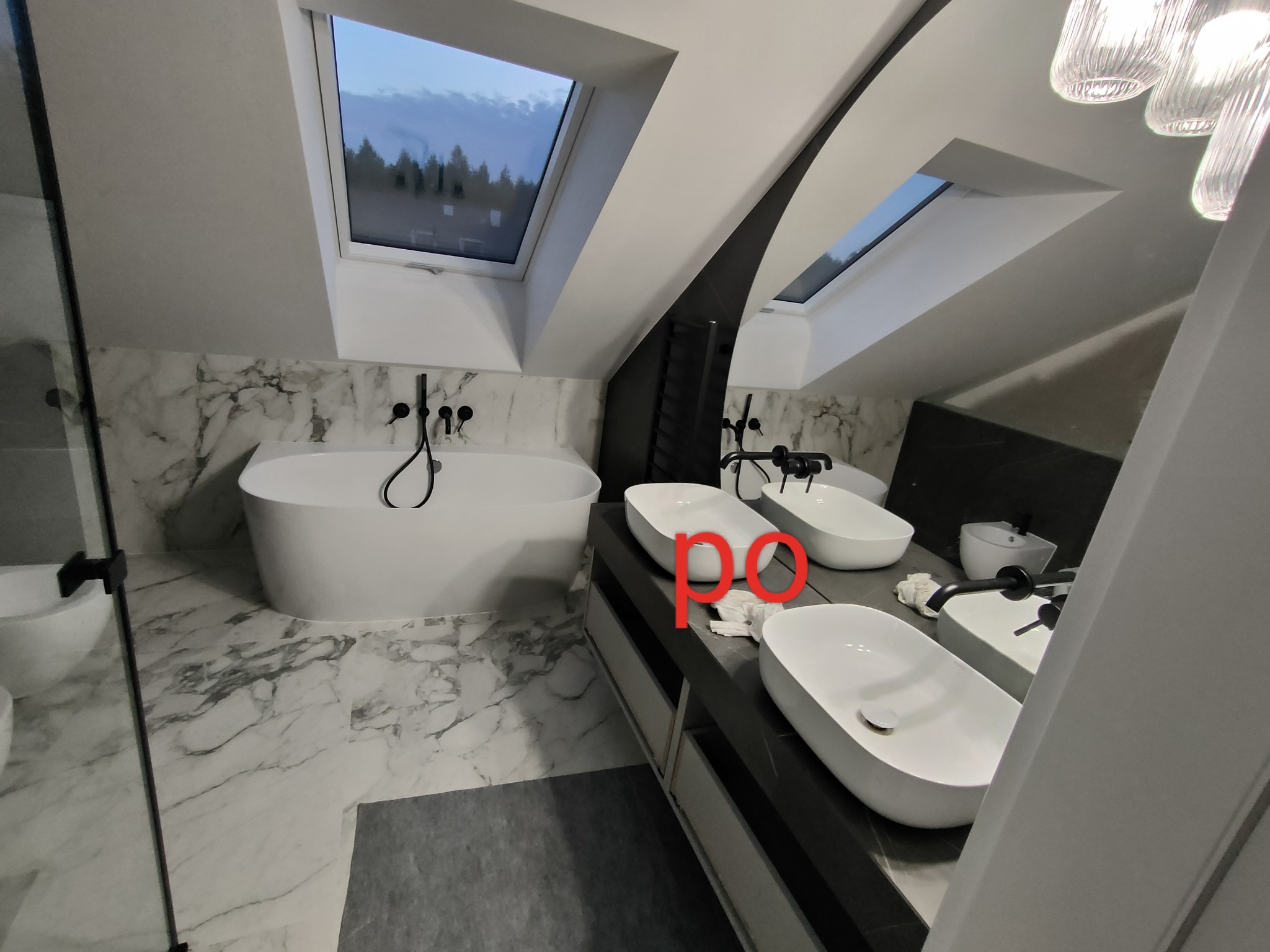 Nowoczesna łazienka na poddaszu z marmurowymi płytkami, wolnostojącą wanną, trzema umywalkami nablatowymi, czarną armaturą i oknami dachowymi z widokiem na las o zmierzchu.