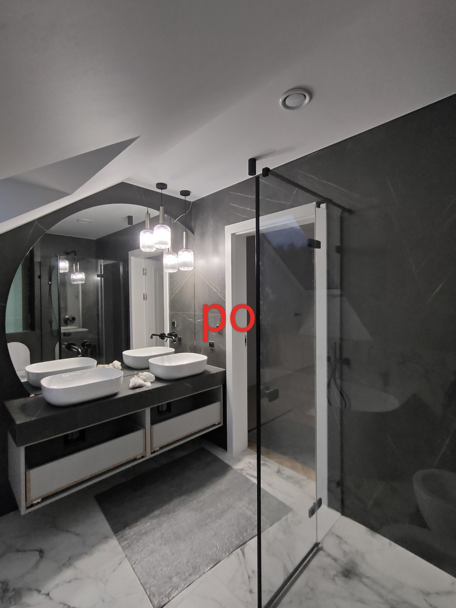 Nowoczesna łazienka z dwoma umywalkami nablatowymi, czarnymi ścianami z marmurowym wzorem, szklaną kabiną prysznicową i designerskimi lampami wiszącymi.