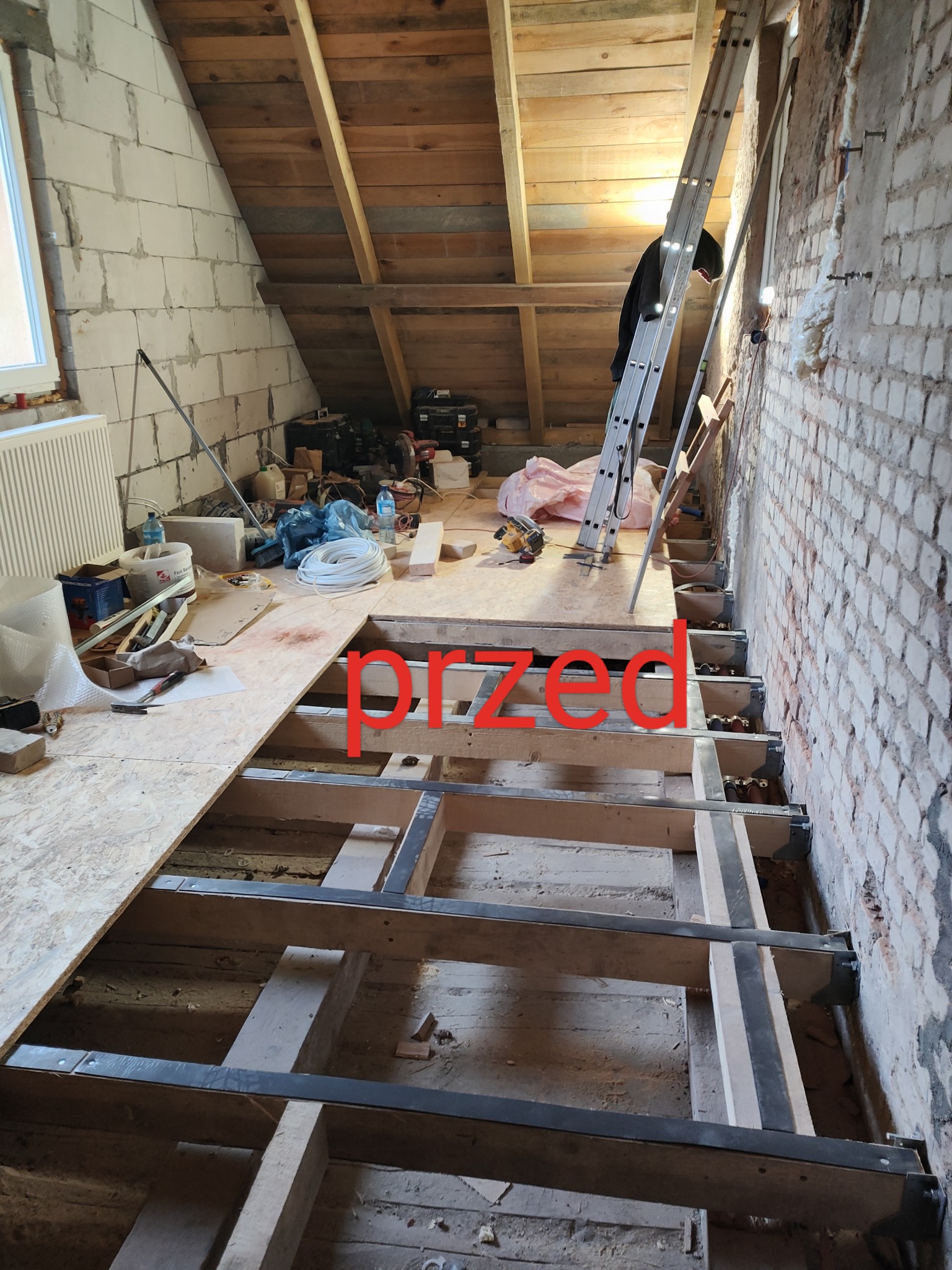 Adaptacja poddasza: widok na drewniany szkielet podłogi z częściowo ułożoną płytą OSB, narzędzia i materiały budowlane porozrzucane, drabina oparta o ceglaną ścianę.