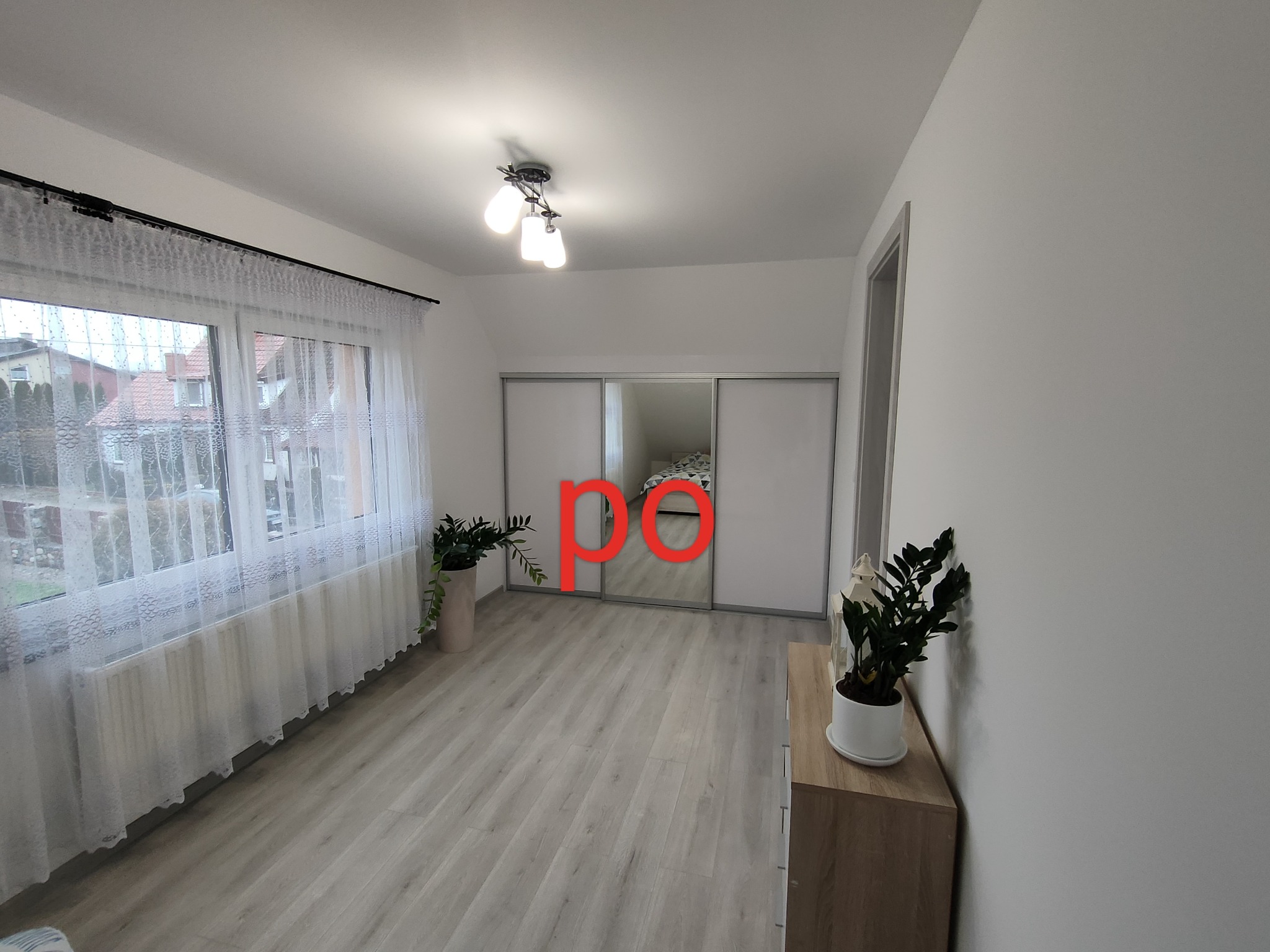 Jasne wnętrze pokoju z panelami podłogowymi, szafą z przesuwnymi drzwiami z lustrem i roślinami doniczkowymi na parapecie oraz komodzie.