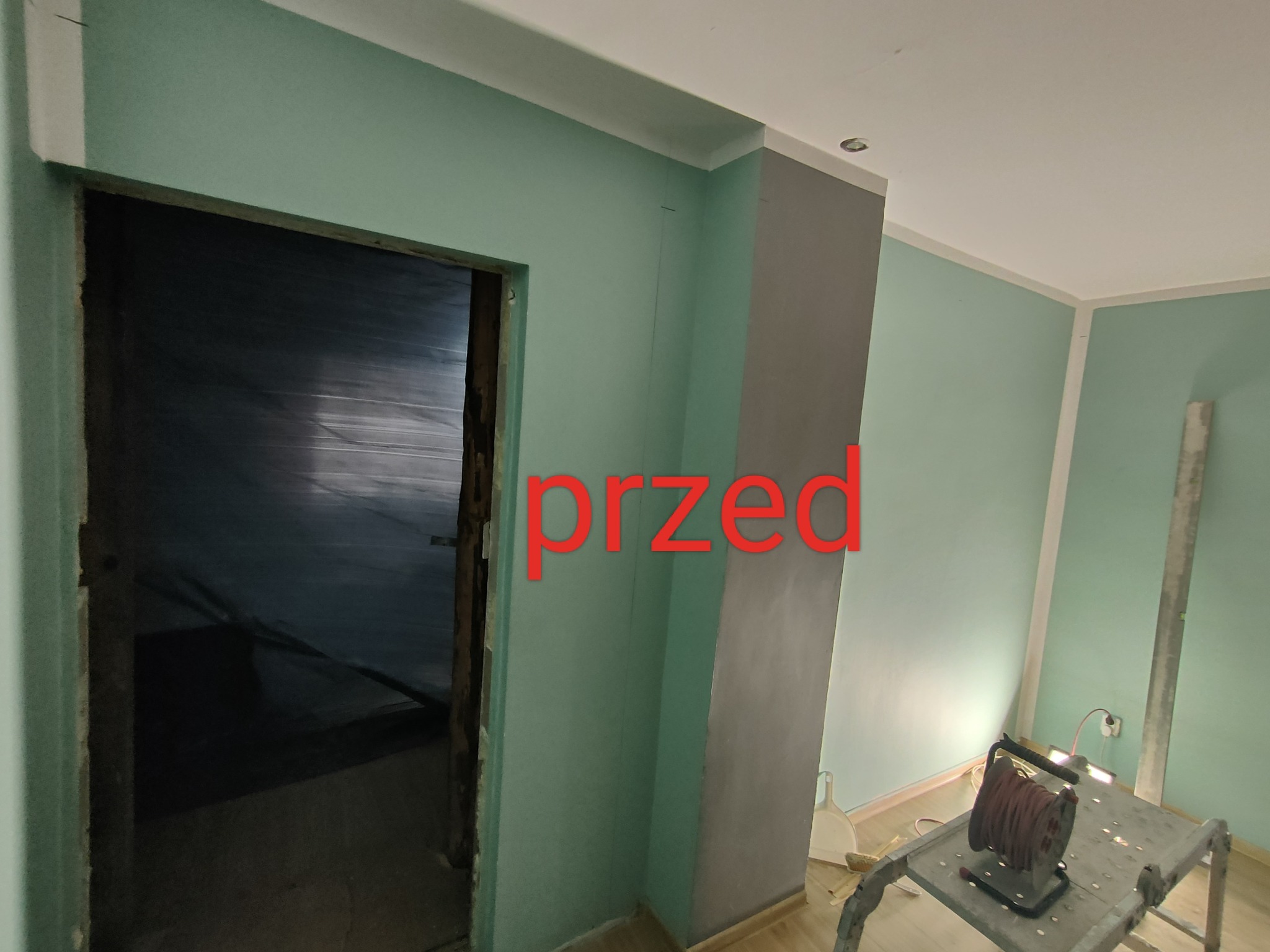 Wnętrze pokoju w trakcie przygotowań do remontu, widoczne ściany w odcieniach zieleni, surowa framuga drzwi, szpachlowana kolumna oraz rozłożony przedłużacz na podeście.