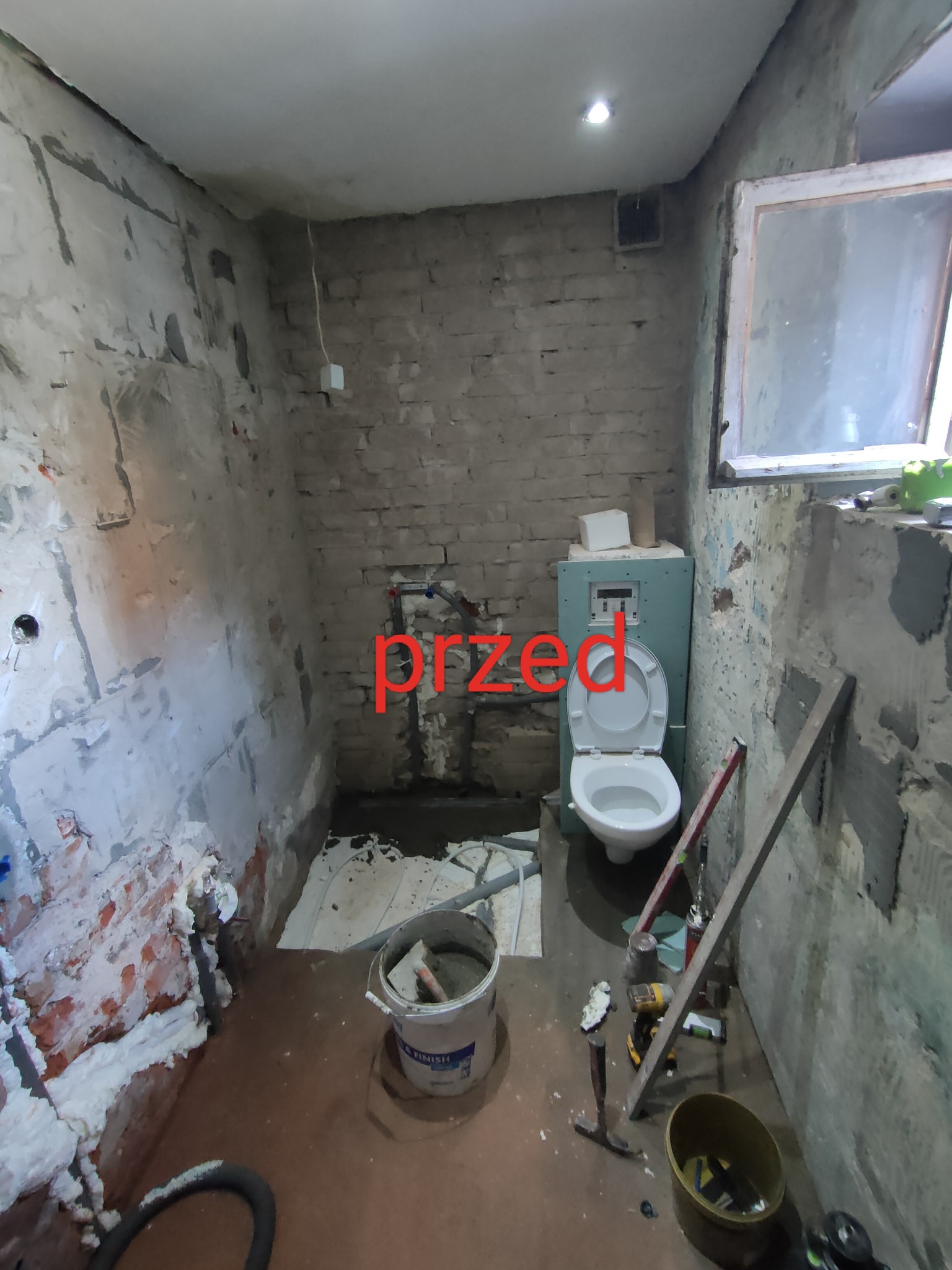 Łazienka przed remontem: surowe ściany z cegły i resztkami tynku, widoczne rury, częściowo zamontowana toaleta, narzędzia i wiadro z zaprawą na podłodze.