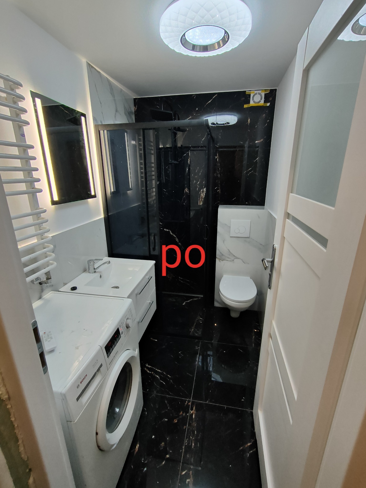 Nowoczesna łazienka z czarnymi marmurowymi płytkami, białą armaturą i pralką Bosch, oświetlona lampą sufitową z efektem kryształu.