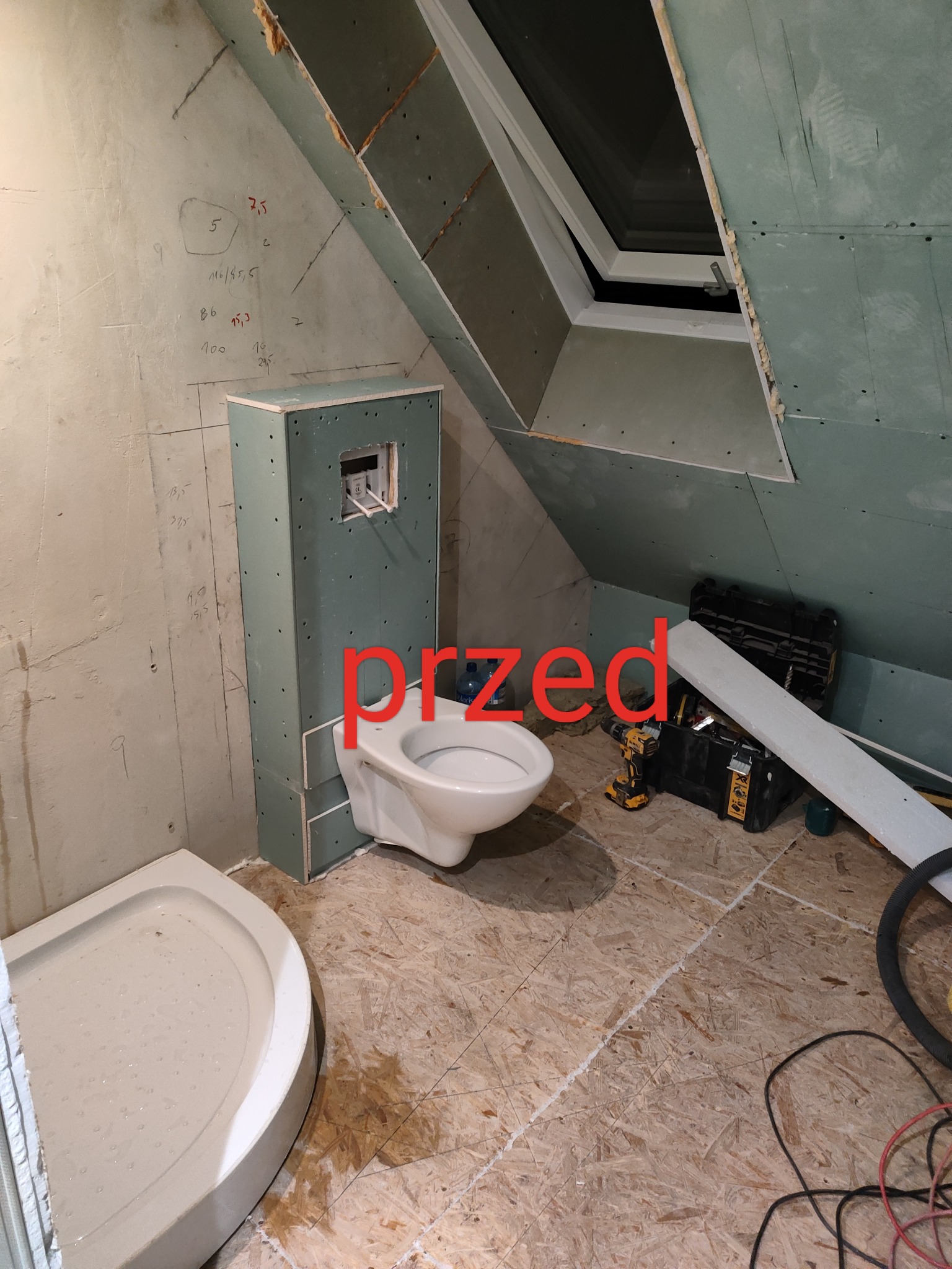 Łazienka na poddaszu w trakcie remontu, widoczna obudowa stelaża WC z płyt kartonowo-gipsowych, brodzik, narzędzia i skrzynka narzędziowa na podłodze z płyt OSB, ściany częściowo wykończone płytami...