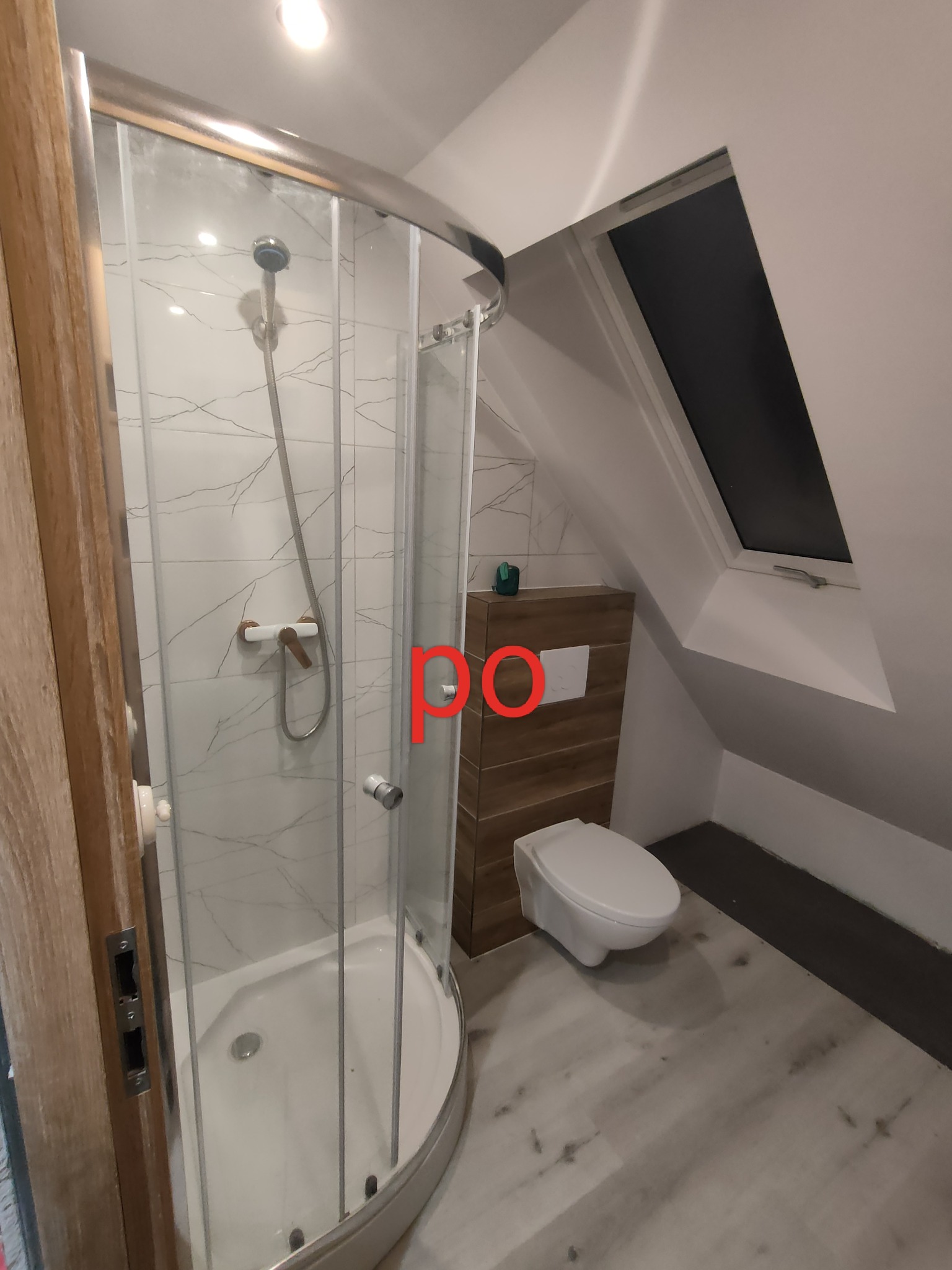 Wykończona łazienka na poddaszu ze skośnym oknem, kabina prysznicowa z chromowanymi ramami, toaleta podwieszana i drewnopodobne płytki na ścianie.