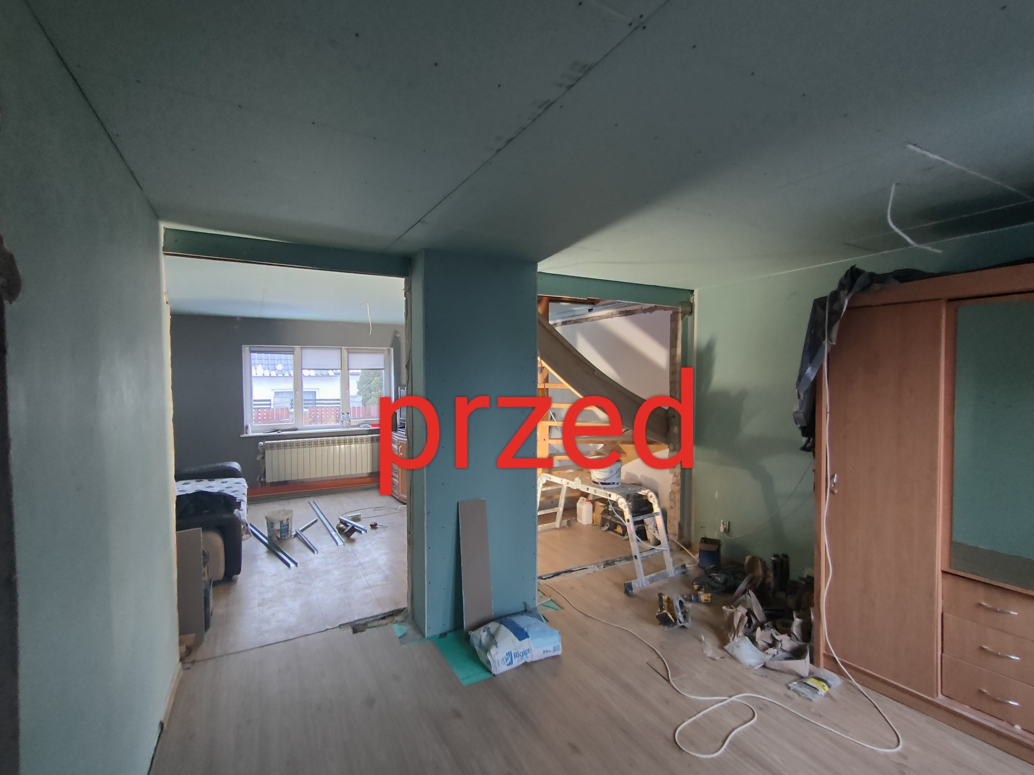 Wnętrze mieszkania w trakcie prac remontowych: widoczne płyty gipsowo-kartonowe na ścianach i suficie, narzędzia pracy, drabina oraz materiały budowlane na podłodze. Zdjęcie z oznaczeniem 'przed'...