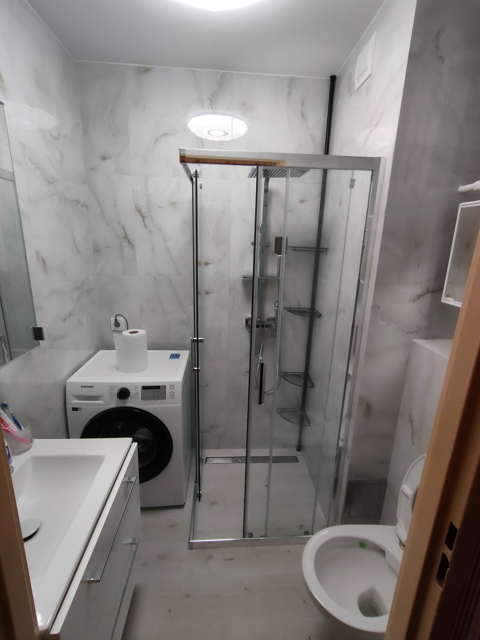 Nowoczesna łazienka z kabiną prysznicową, umywalką z szafką, pralką Samsung i toaletą, wykończona marmurowymi płytkami.