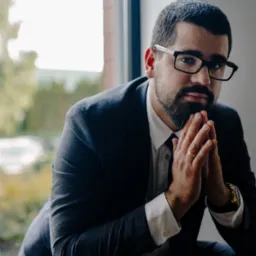 Mężczyzna w garniturze i okularach siedzi przy oknie z rękami złożonymi w geście skupienia, widoczny złoty zegarek i obrączka.