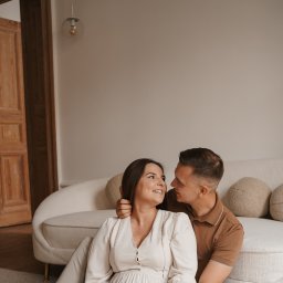 Klaudia Makowska-Kuźma - Uśmiechnięta para siedzi na podłodze w jasnym wnętrzu, ona w kremowej sukience, on w brązowej koszuli polo, patrzą na siebie z czułością, w tle kremowa sofa i drewniane drzwi.
