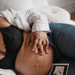 Klaudia Makowska-Kuźma - Brzuch kobiety w ciąży, dłonie partnera z obrączką, zdjęcie USG dziecka w rogu kadru, biała koszula i dżinsy.