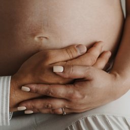 Klaudia Makowska-Kuźma - Brzuch kobiety w ciąży, dłonie partnera z obrączką obejmujące ciążowy brzuszek, beżowe paznokcie, sukienka w neutralnym odcieniu.