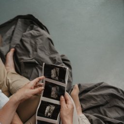 Klaudia Makowska-Kuźma - Rodzice siedzą na łóżku, trzymając w rękach serię wydruków USG dziecka, z widocznymi szczegółami twarzy nienarodzonego dziecka.