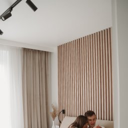 Klaudia Makowska-Kuźma - Młoda para w objęciach na łóżku z drewnianym panelem ściennym w tle, oświetlenie punktowe na suficie i zasłony w odcieniach beżu.