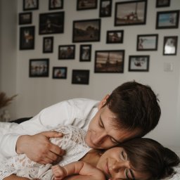 Klaudia Makowska-Kuźma - Uśmiechnięta para leży blisko, mężczyzna obejmuje kobietę ubraną w koronkową bluzkę, w tle ściana z galerią zdjęć w czarnych ramkach.