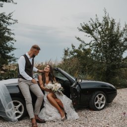 Klaudia Makowska-Kuźma - Para młoda pozuje przy czarnym kabriolecie na tle zieleni, panna młoda trzyma bukiet białych róż, a pan młody opiera się o samochód.