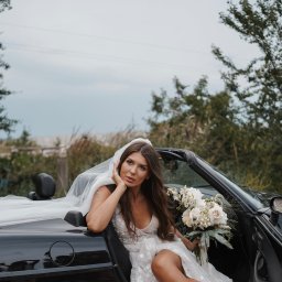 Klaudia Makowska-Kuźma - Panna młoda w sukni ślubnej z bukietem, siedząca w czarnym kabriolecie na tle zieleni i jasnego nieba.