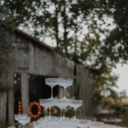 Klaudia Makowska-Kuźma - Piramida z kryształowych kieliszków na tle drewnianej stodoły z podświetlonym napisem LOVE, otoczonej zielenią.