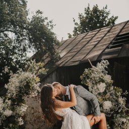 Klaudia Makowska-Kuźma - Para młoda podczas pocałunku w plenerze, panna młoda w koronkowej sukni z tatuażem na ramieniu, pan młody w szarej marynarce w kratę, rustykalna stodoła w tle, dekoracja z białych kwiatów.