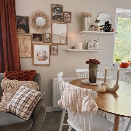 Klaudia Makowska-Kuźma - Przytulne wnętrze pokoju z galerią zdjęć na ścianie, drewnianym stołem, białymi krzesłami i dekoracjami jesiennymi, takimi jak dynia i świece.